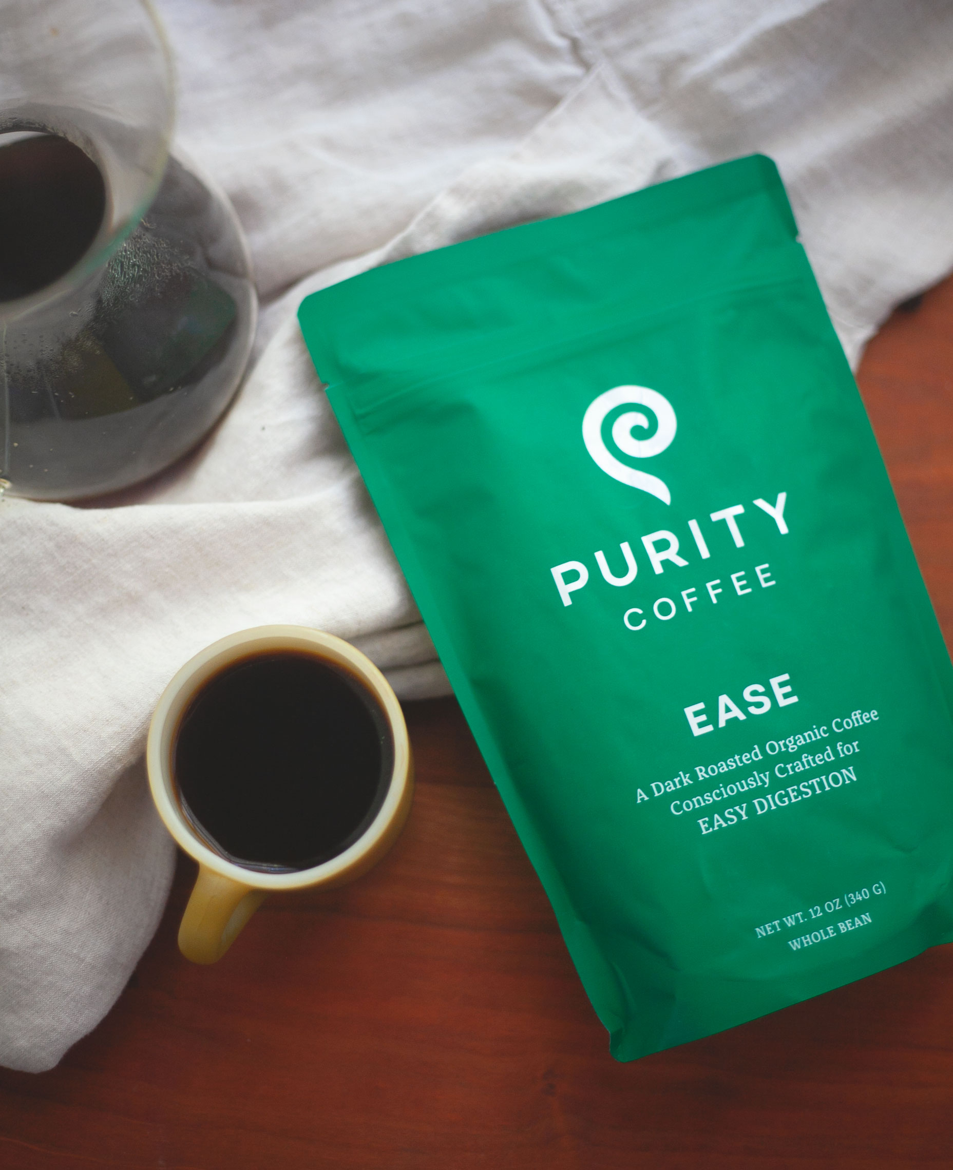 A vibrant green bag of Purity Coffee labeled "Ease" sits next to a cup of freshly brewed dark coffee, with a glass coffee pot nearby, all on a light-colored cloth background.